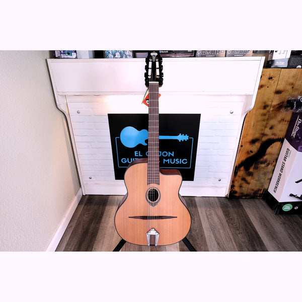 A Lâg SL100 Petite Bouche Gypsy Jazz Guitar acoustic guitar with a natural wood body and black headstock stands on a stand in front of a the front counter of EL Cajon Guitars and Music, with the logo showing at the front counter behind the guitar in the stand.