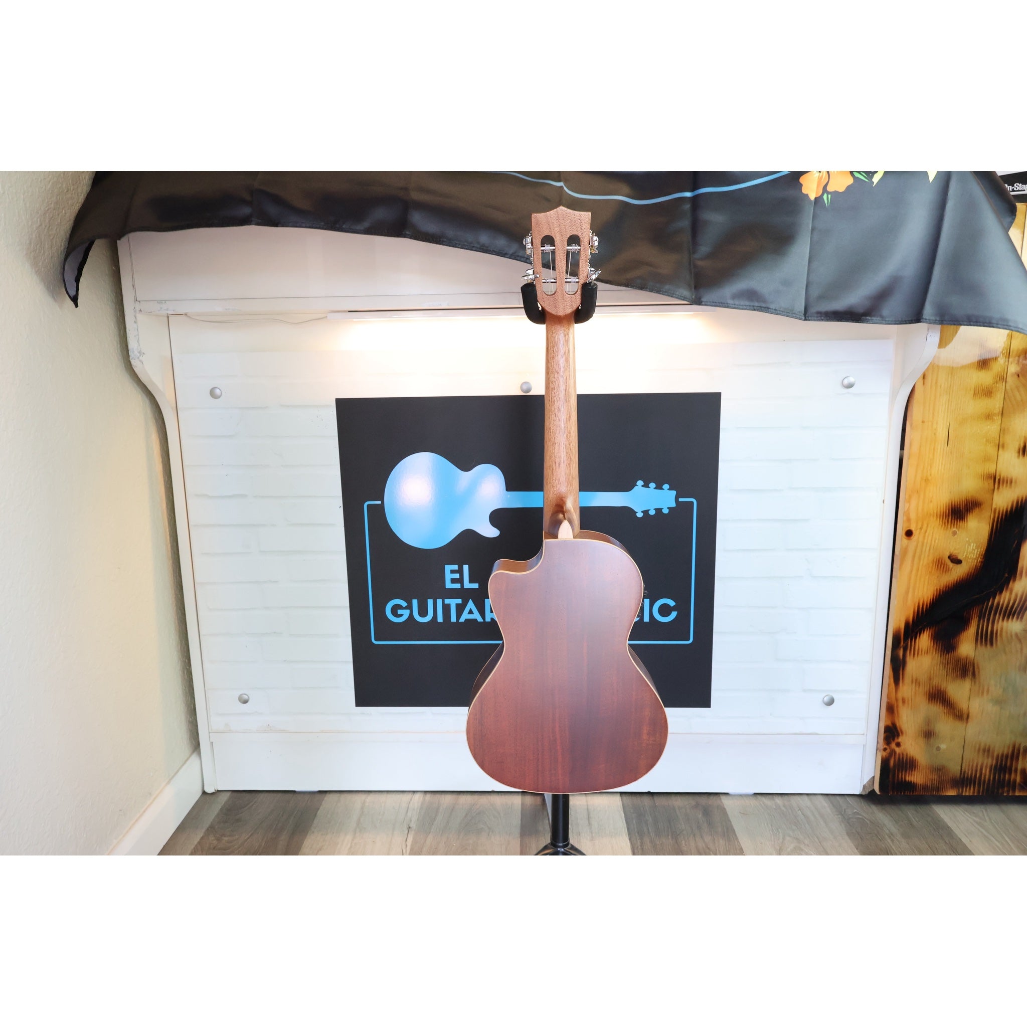 The back of a Kala KA-SA-TE-C All-Solid Acacia Tenor Cutaway Ukulele with Fishman Electronics ukulele standing upright on a display stand in from of El Cajon Gutiars & Music Logo at the shop,.