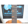 Close-up of a Kala KA-WF-AMHG-T All Solid Mahogany Waterfall Tenor Ukulele with a warm brown wooden body and dark fretboard, standing in front of a white brick wall and a black sign that reads El Cajon Guitars & Music.