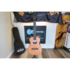 A Kala KA-WF-AMHG-T All Solid Mahogany Waterfall Tenor Ukulele at ECGMusic with a natural wood body and black headstock is displayed on a stand in front of a white brick wall, with its black gig bag leaning to the left.