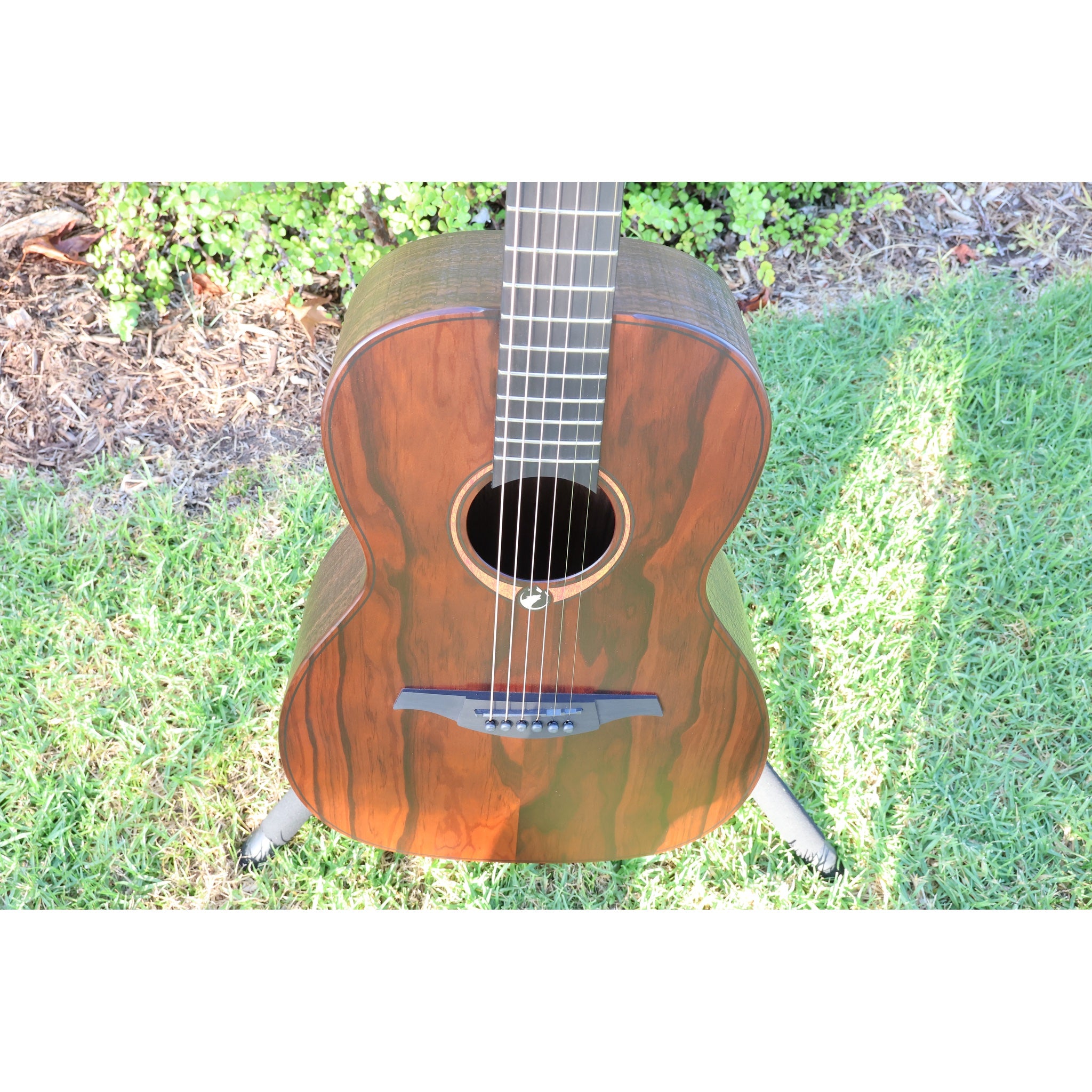 Front top view of a Lâg Sauvage PE Parlor Acoustic-Electric Guitar body with a rich brown grain pattern, showing the sound hole, strings, and bridge, set against a background of grass and mulch.