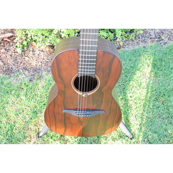 Front top view of a Lâg Sauvage PE Parlor Acoustic-Electric Guitar body with a rich brown grain pattern, showing the sound hole, strings, and bridge, set against a background of grass and mulch.