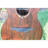 Close-up of a Lâg Sauvage PE Parlor Acoustic-Electric Guitar body with a rich brown grain pattern, showing the sound hole, strings, and bridge, set against a background of grass and mulch.