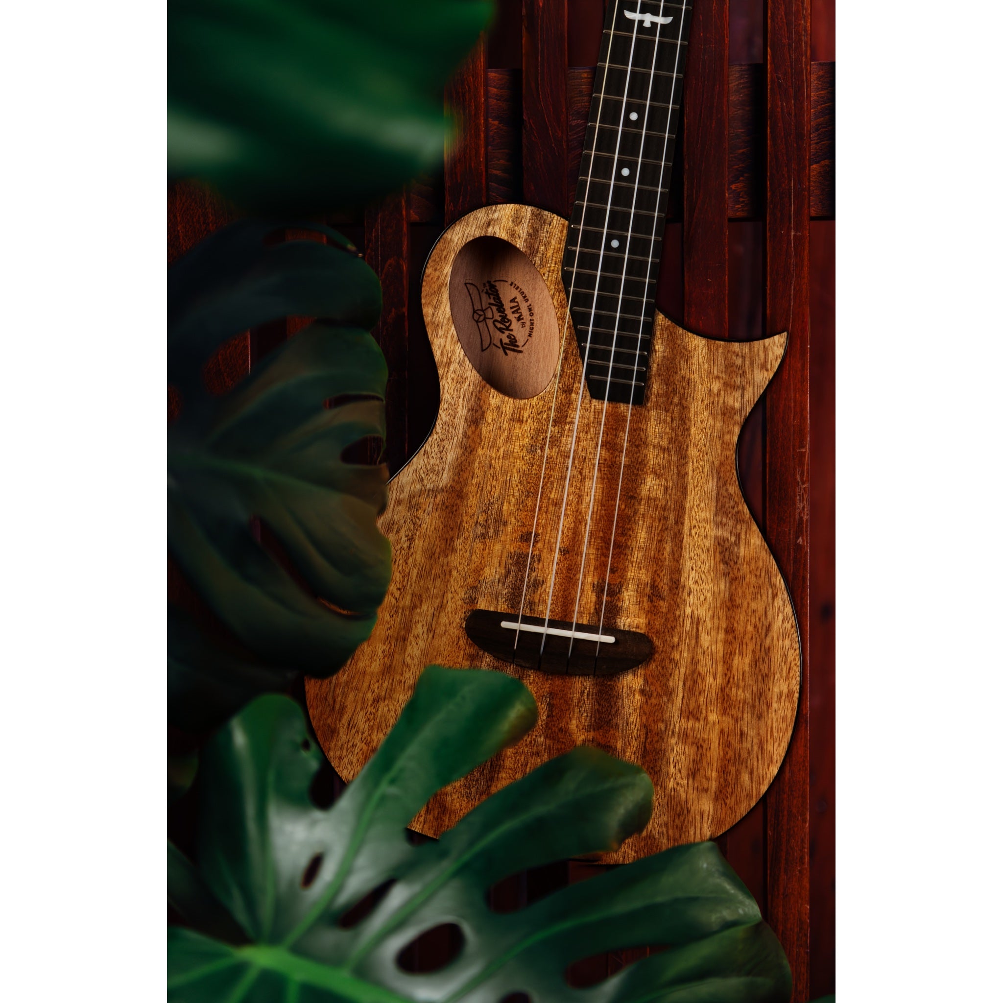 A Kala Revelator Night Owl Mango Tenor Acoustic-Electric Ukulele with a natural wood grain body and dark neck, styled with green tropical leaves against a dark wooden background.