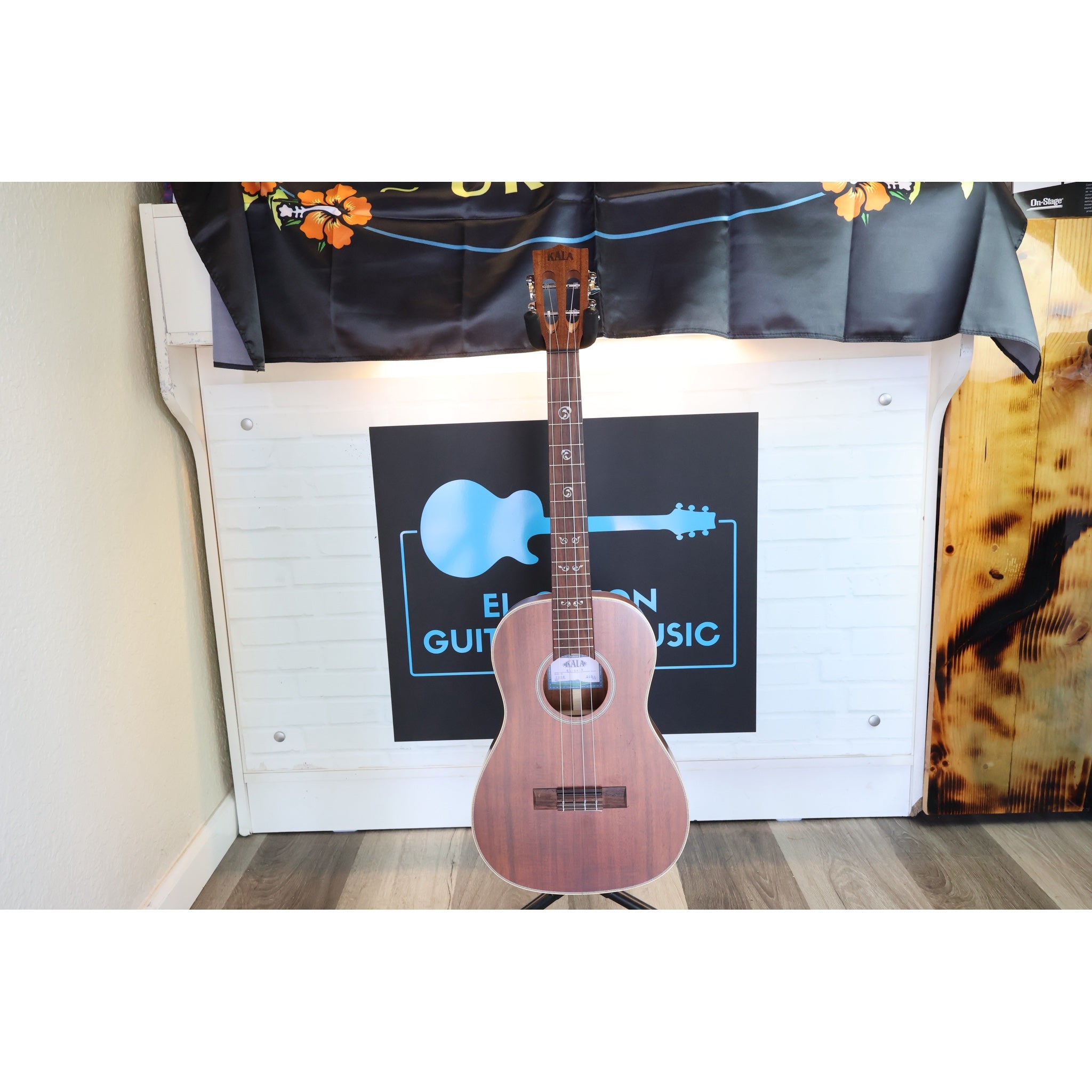 A A Kala KA-SA-B All-Solid Acacia Baritone Ukulele with a reddish-brown finish stands on a stand in front of a white paneled wall with a black sign that reads 'Eaton Guitar Music.