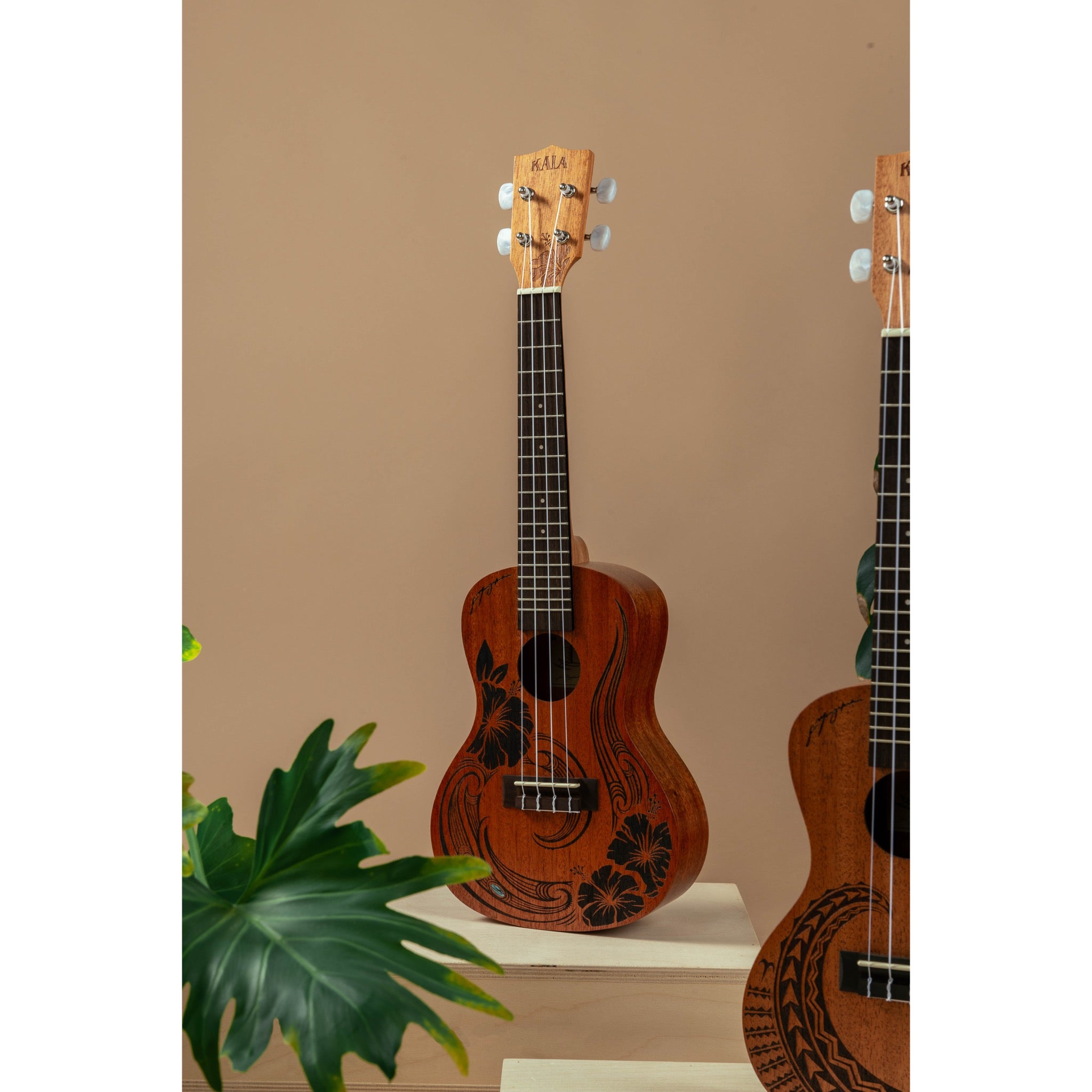 A Kala Unity Mahogany Tenor Ukulele w/ Bag - KA-UNITY-T_W/BAG with black floral inlay stands on a white pedestal against a beige wall, with another ukulele partially visible on the right and a green plant leaf in the lower left.