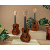 Kala Unity Mahogany Tenor Ukulele w/ Bag - KA-UNITY-T_W/BAG with decorative black inlay patterns are displayed on light wood pedestals against a warm beige background, surrounded by green houseplants with other ukuleles in the series.