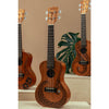Kala Unity Mahogany Tenor Ukulele w/ Bag - KA-UNITY-T_W/BAG  with dark floral and tribal-style inlays are displayed on light wood steps against a warm beige background, with a green monstera leaf prop in the center with other ukuleles in the series.
