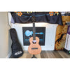 A Kala KA-WF-AMHG-T All Solid Mahogany Waterfall Tenor Ukulele at ECG Music with a natural wood finish stands on a stand in front of a garage door, with its black gig bag to the left and a decorative Kala banner above.