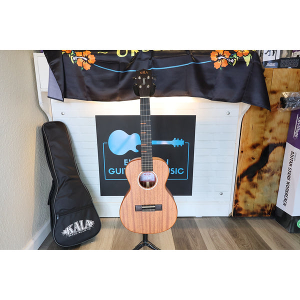 A Kala KA-WF-AMHG-T All Solid Mahogany Waterfall Tenor Ukulele at ECG Music with a natural wood finish stands on a stand in front of a garage door, with its black gig bag to the left and a decorative Kala banner above.
