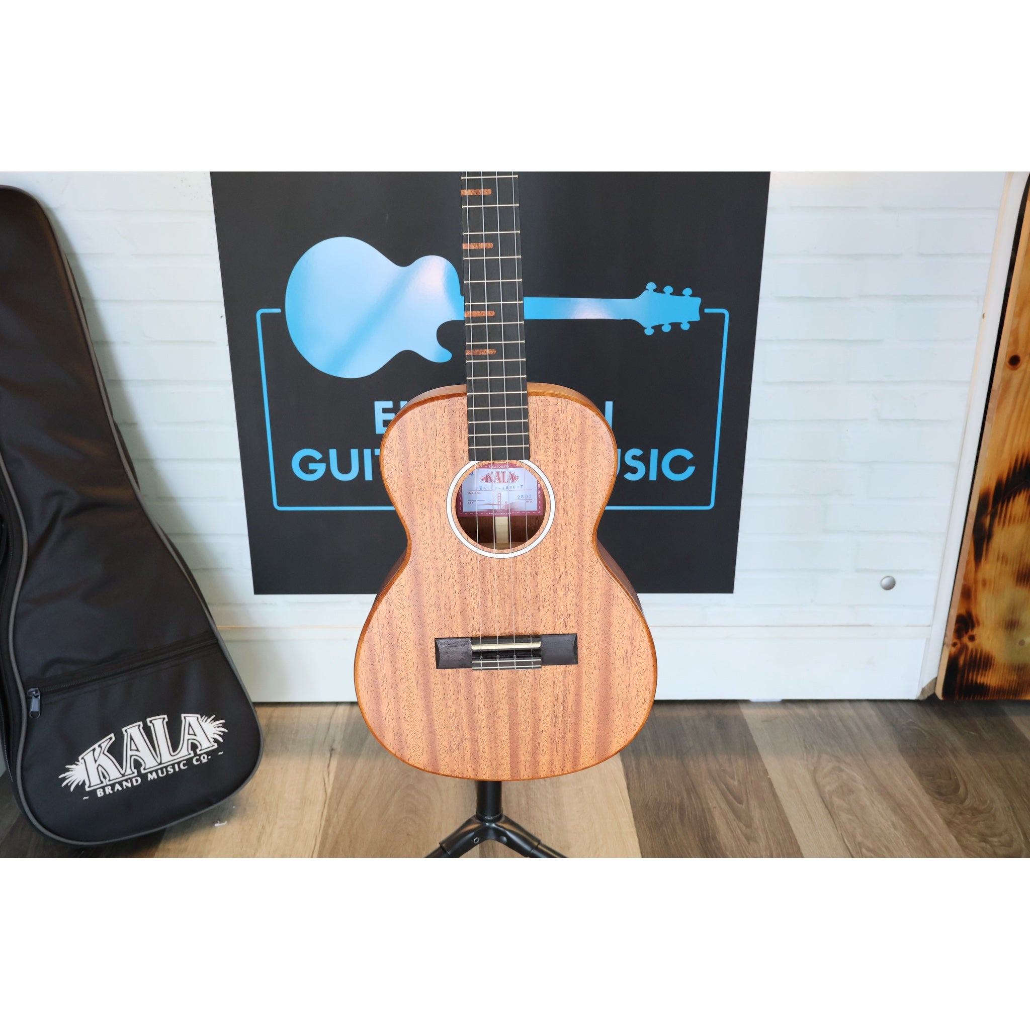 Close-up of a Kala KA-WF-AMHG-T All Solid Mahogany Waterfall Tenor Ukulele at ECGMusic with a natural wood finish stands on a black stand in front of a white brick wall, with a black Kala guitar case on the left ECG Music logo behind it.