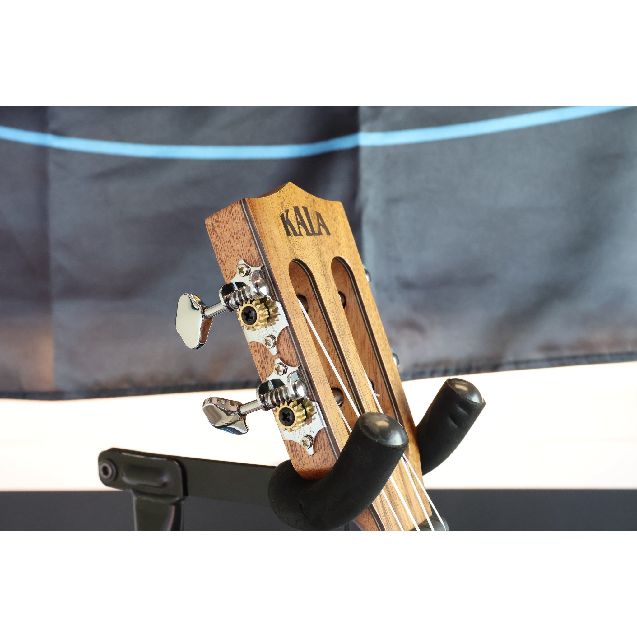 Side headstock Close-up of a A Kala KA-SA-TE-C All-Solid Acacia Tenor Cutaway Ukulele with Fishman Electronics ukulele  mounted in a black guitar stand, with tuning pegs and strings....showing the Kala logo and amazing tuners on this insrument.