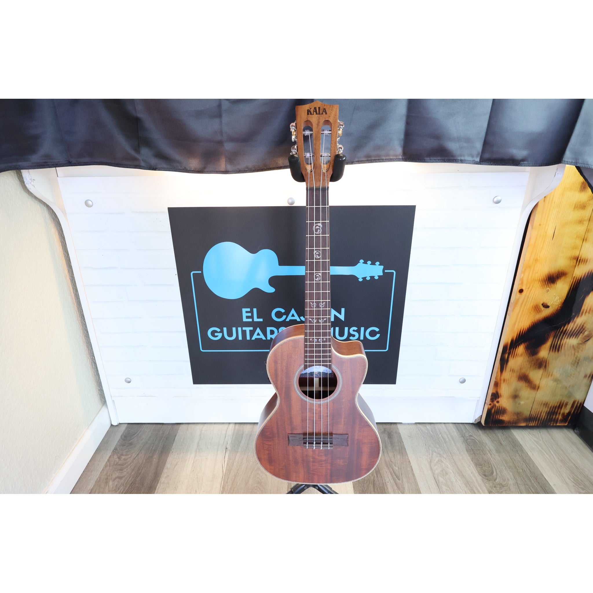 A Kala brand  Close-up of a A Kala KA-SA-TE-C All-Solid Acacia Tenor Cutaway Ukulele with Fishman Electronics ukulele  mounted in a black guitar stand, with tuning pegs and strings visible against the ECGMusic logo.