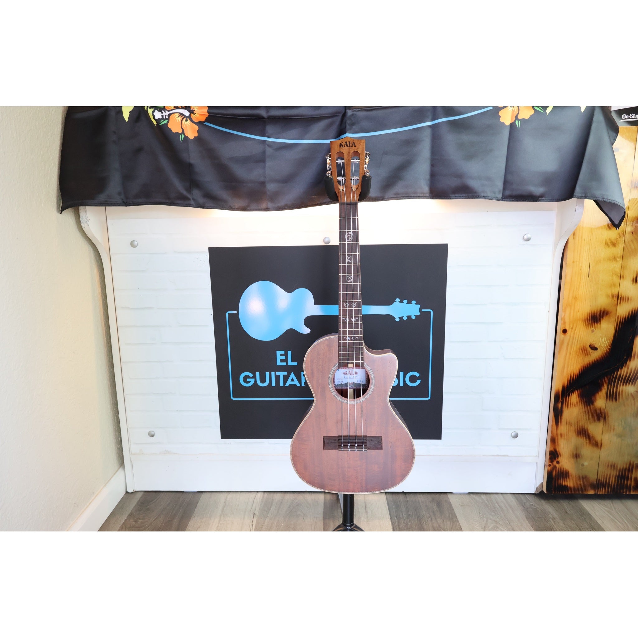 A Kala KA-SA-TE-C All-Solid Acacia Tenor Cutaway Ukulele with Fishman Electronics ukulele standing upright on a display stand in from of El Cajon Gutiars & Music Logo at the shop.
