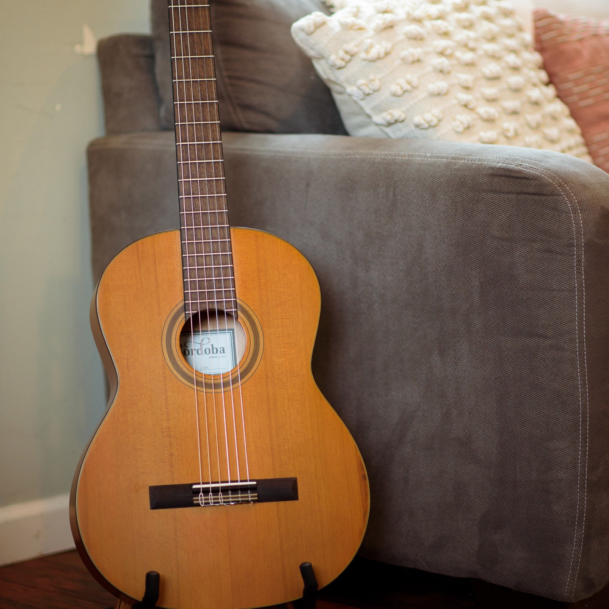 Córdoba C3M Acoustic guitar on a stand against a gray couch with decorative pillows.