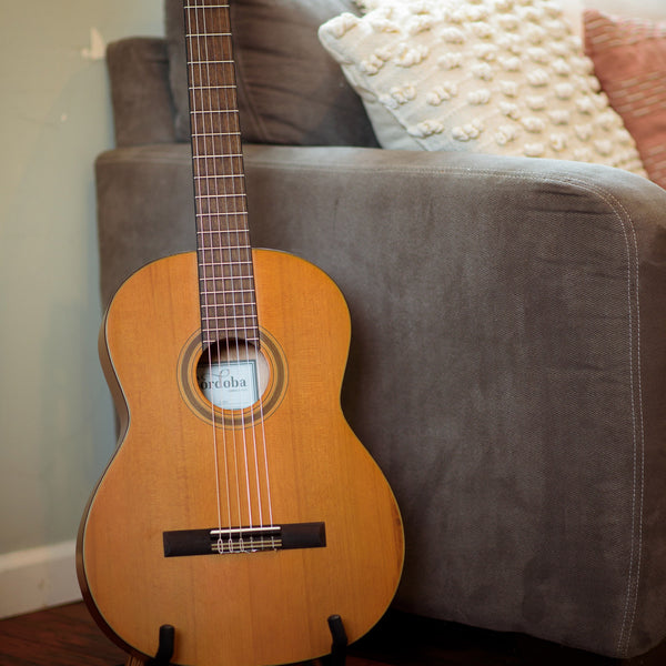 Córdoba C3M Acoustic guitar on a stand against a gray couch with decorative pillows.