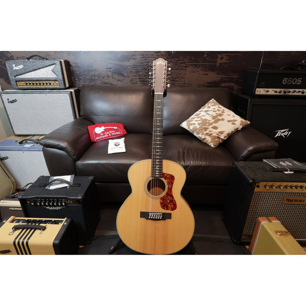 Acoustic guitar on a stand in front of a brown leather couch with amplifiers and a pillow in the background.