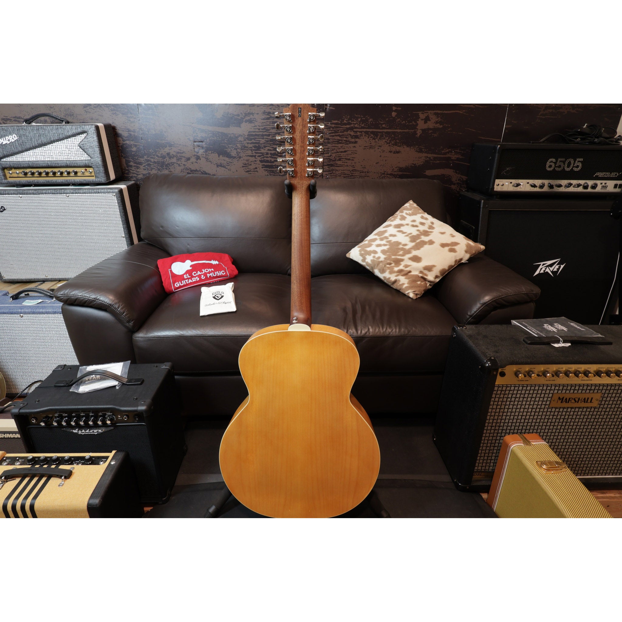 Acoustic guitar on a stand in front of a brown leather couch with amplifiers and speakers around.