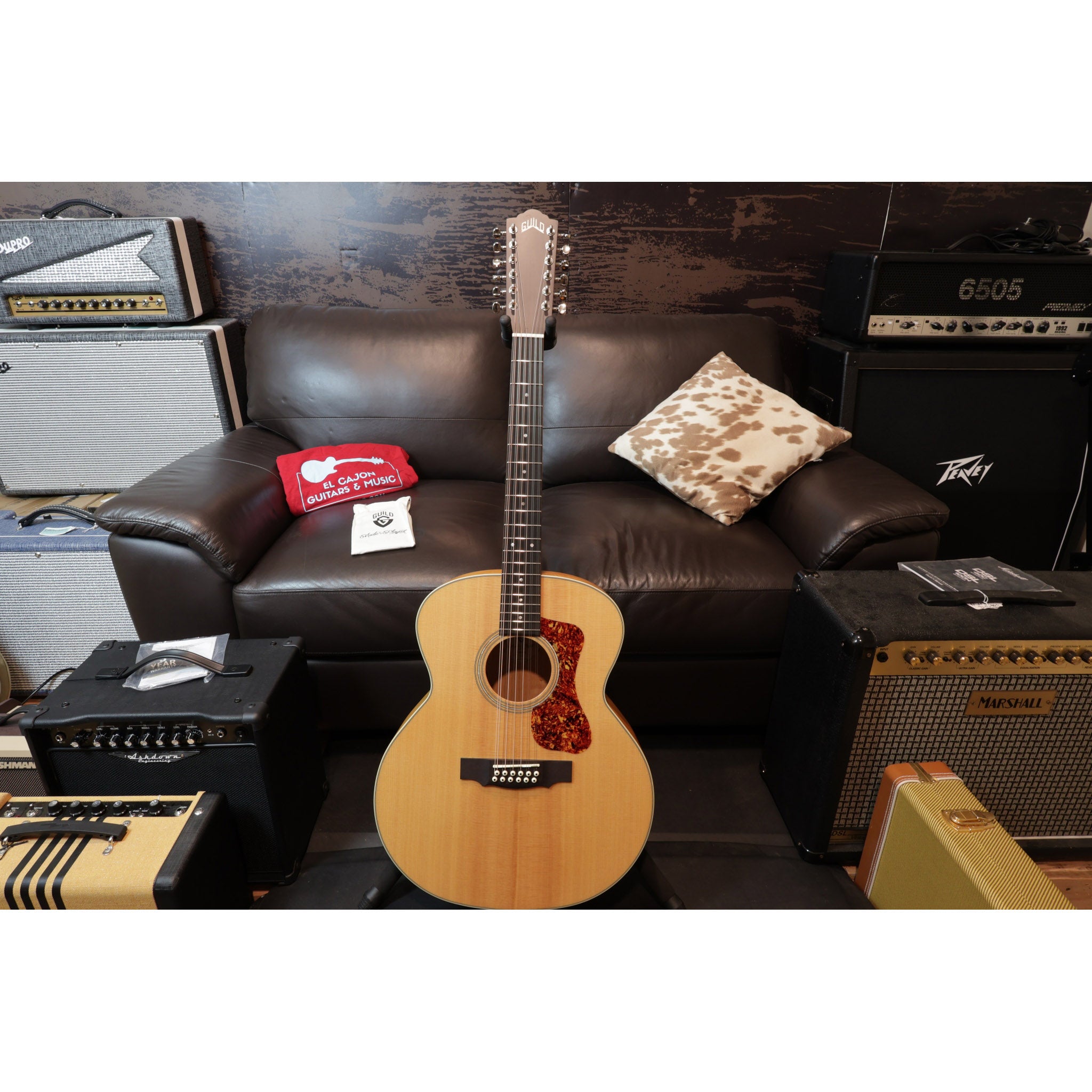 Acoustic guitar on a stand in front of a brown leather couch with amplifiers and equipment around.