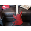 Red electric guitar on a stand with a brown leather couch and a red pillow in the background.