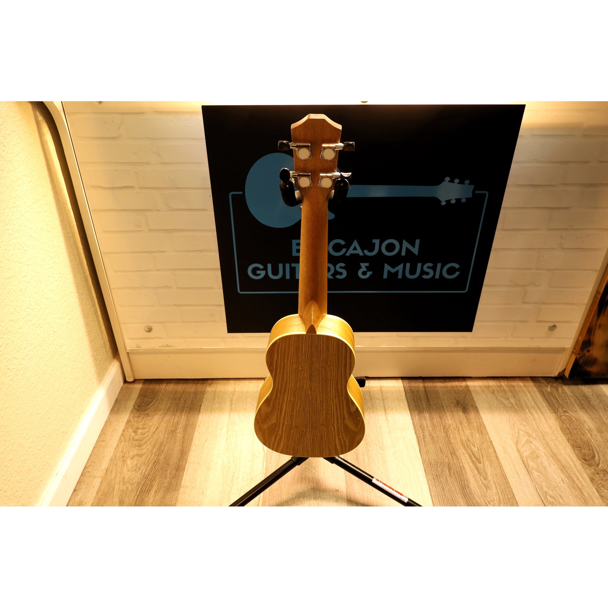 Wooden ukulele on a stand with a blurred background featuring 'Bacajon Guitars & Music'.