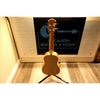 Wooden ukulele on a stand with a blurred background featuring 'Bacajon Guitars & Music'.