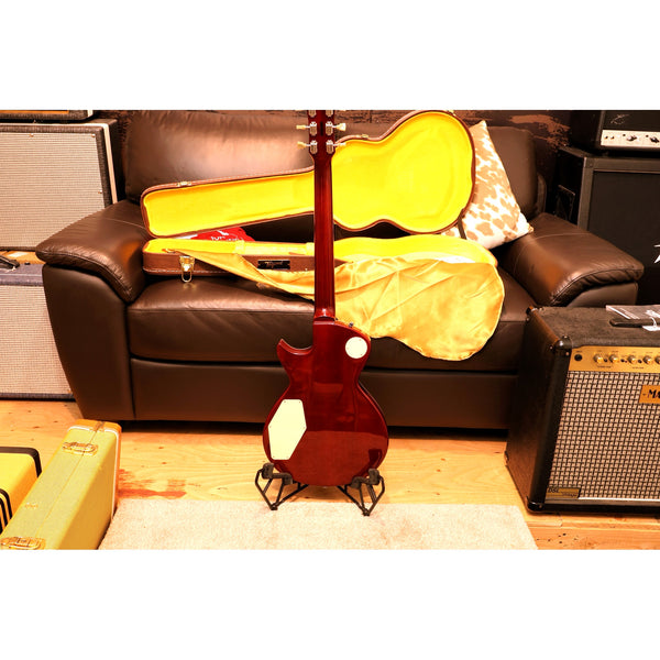 Guitar in a yellow case on a stand in front of a brown couch with an amplifier to the side.