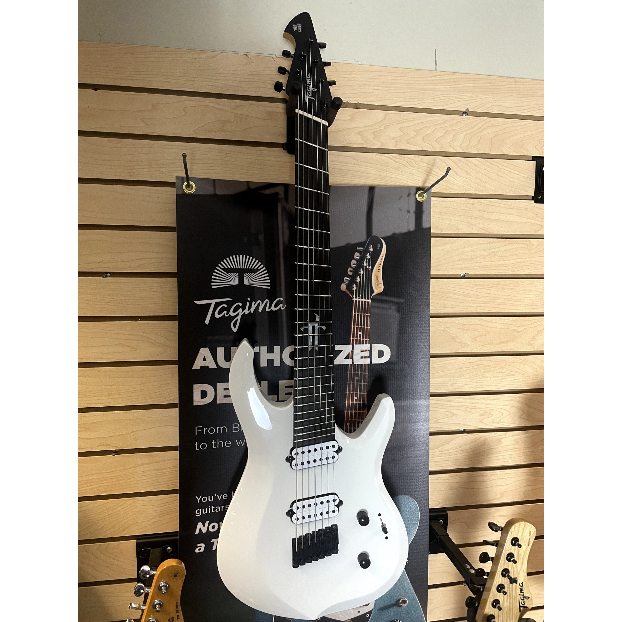 White electric guitar leaning against a wooden wall with a Tagima banner.