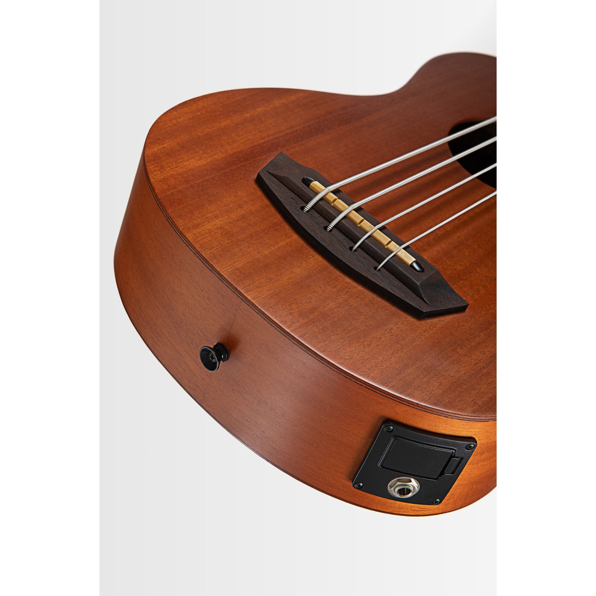 Close-up of a Kala Nomad Acoustic-Electric U•BASS® - UBASS-NOMAD-FS wooden electric ukulele body showing the warm brown wood grain, black bridge, strings, and a black input jack on a light gray background.