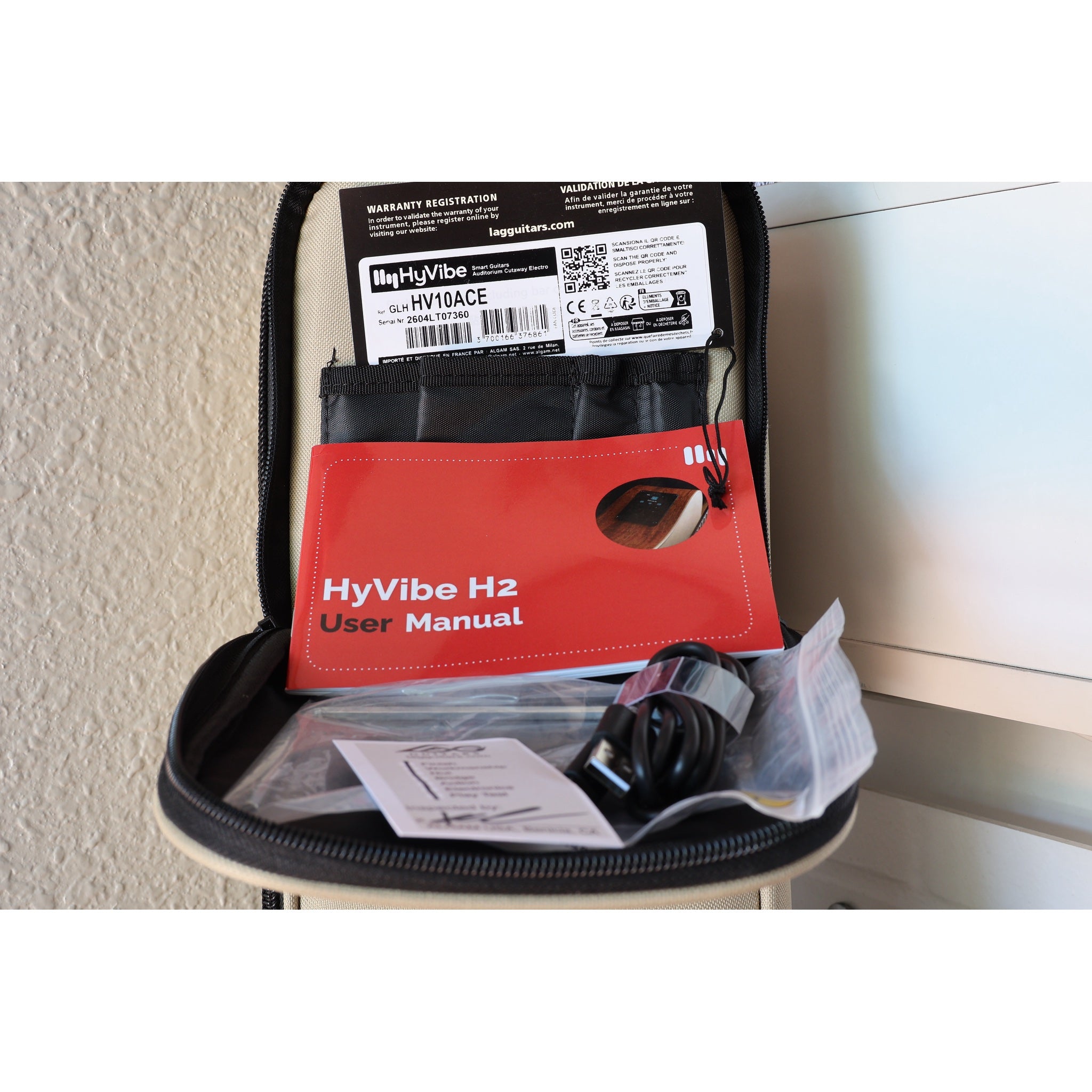 Close up of the Upper case pocket contents for this Lâg HyVibe 10 HV10ACE showing the Warranty Registration Card, HyVibe H2 User Manual, Truss rod adjustment tool, and USB charging cable for this guitar with Serial Number 2604LT07360.