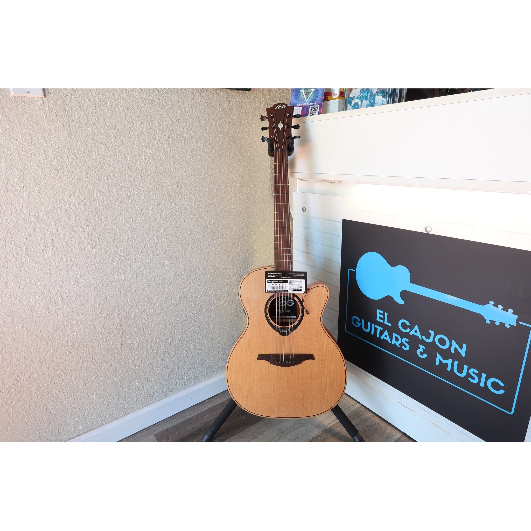 Full guitar view in the stand of this Lâg HyVibe 10 HV10ACE with Warranty Registration Card showing Serial Number 2604LT07360 and UPC hung on the guitar at the strings on the neck at the body, with the soundhole and sticker showing beneath it.
