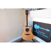 Full guitar view in the stand of this Lâg HyVibe 10 HV10ACE with Warranty Registration Card showing Serial Number 2604LT07360 and UPC hung on the guitar at the strings on the neck at the body, with the soundhole and sticker showing beneath it.