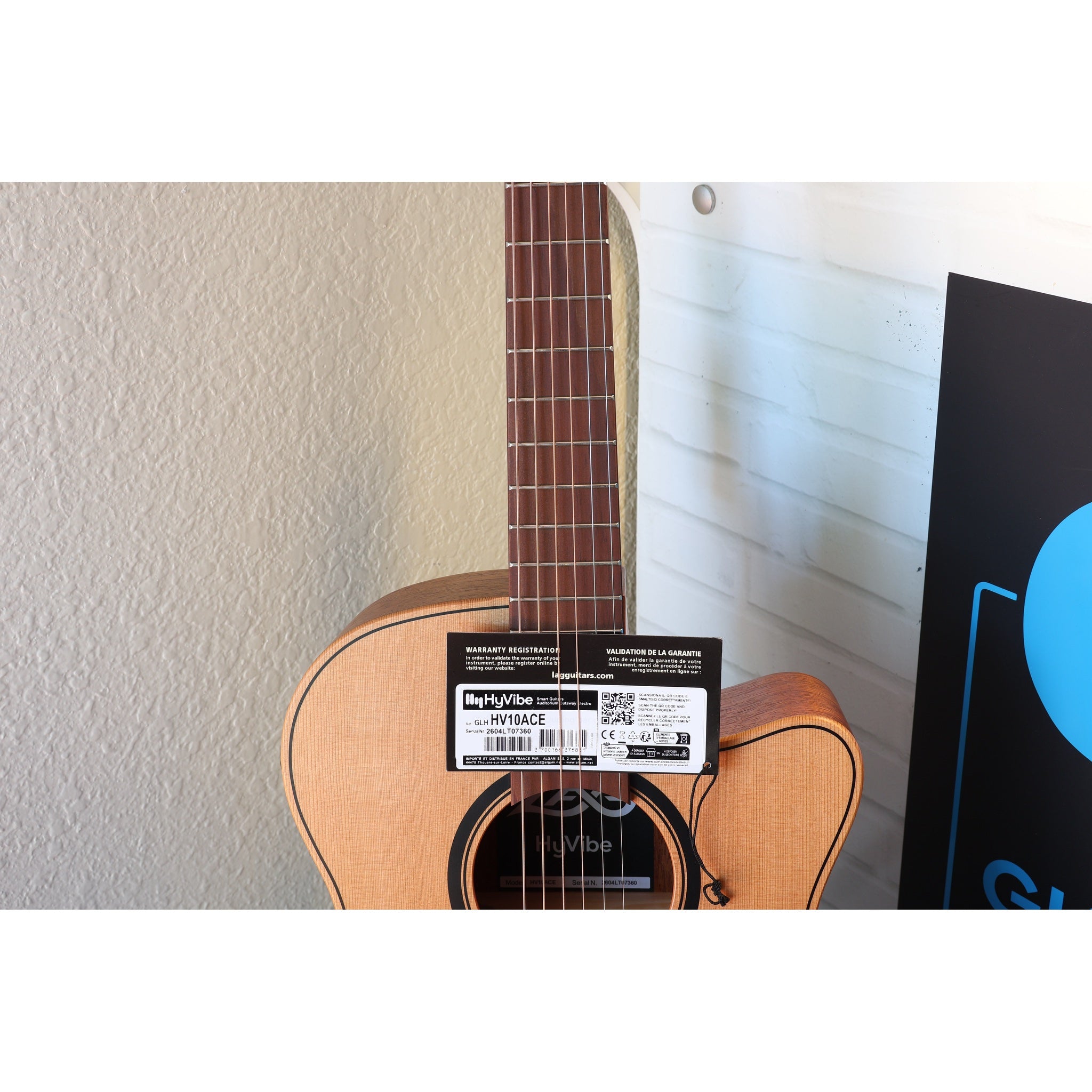 This Lâg HyVibe 10 HV10ACE with Warranty Registration Card showing Serial Number 2604LT07360 and UPC hung on the guitar at the strings on the neck at the body, with the soundhole and sticker showing beneath it.