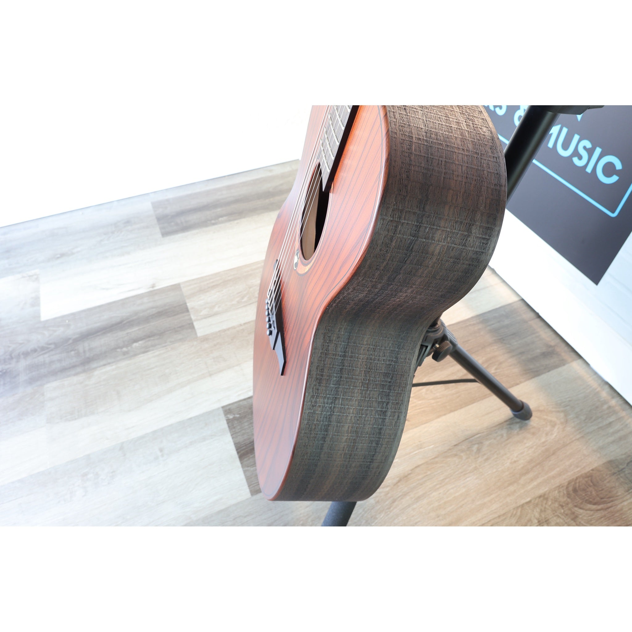 Close-up of an acoustic guitar body on a stand, showing a reddish-brown wood finish and dark gray back, photographed indoors on a light wood floor.