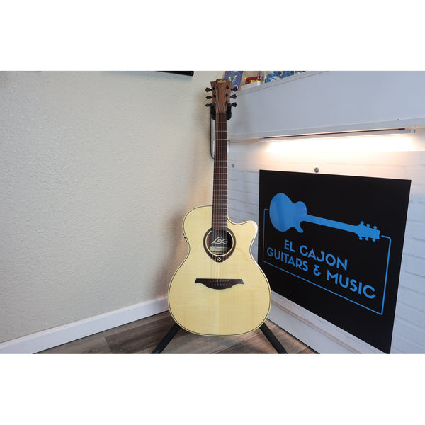 Lâg Tramontane T88ACE Auditorium Cutaway Acoustic-Electric Guitar with a natural light wood finish stands on a black stand in a room corner next to a black sign with a blue guitar graphic and the text 'EL CAJON GUITARS & MUSIC'.