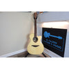 A Lâg Tramontane T88ACE Auditorium Cutaway Acoustic-Electric Guitar acoustic guitar with a natural wood finish stands on a stand in a room corner next to a black sign that reads 'EL CAJON GUITARS & MUSIC'.