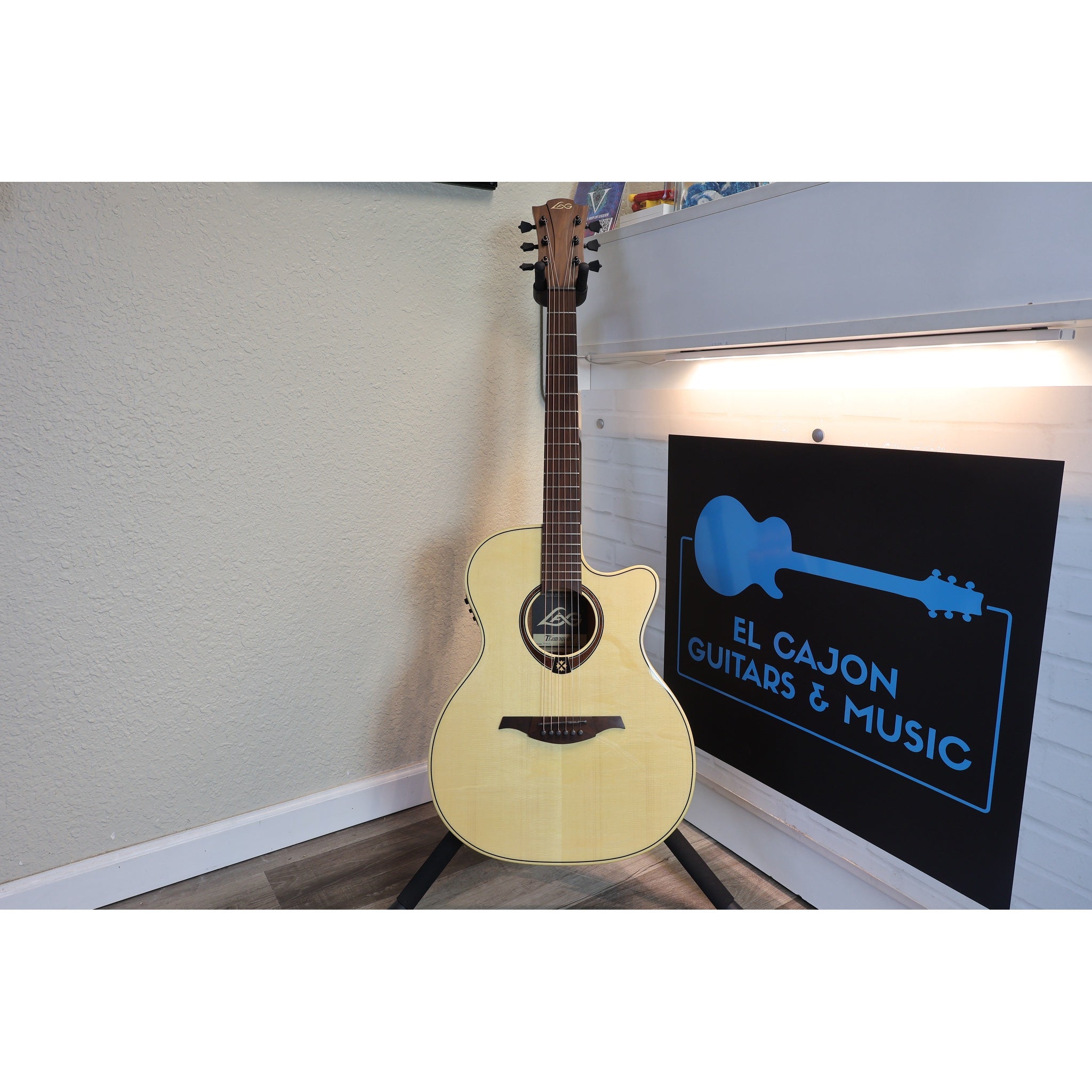 An acoustic guitar with a natural wood finish stands on a stand in a room corner next to a black sign that reads 'EL CAJON GUITARS & MUSIC'.