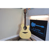 An acoustic guitar with a natural wood finish stands on a stand in a room corner next to a black sign that reads 'EL CAJON GUITARS & MUSIC'.