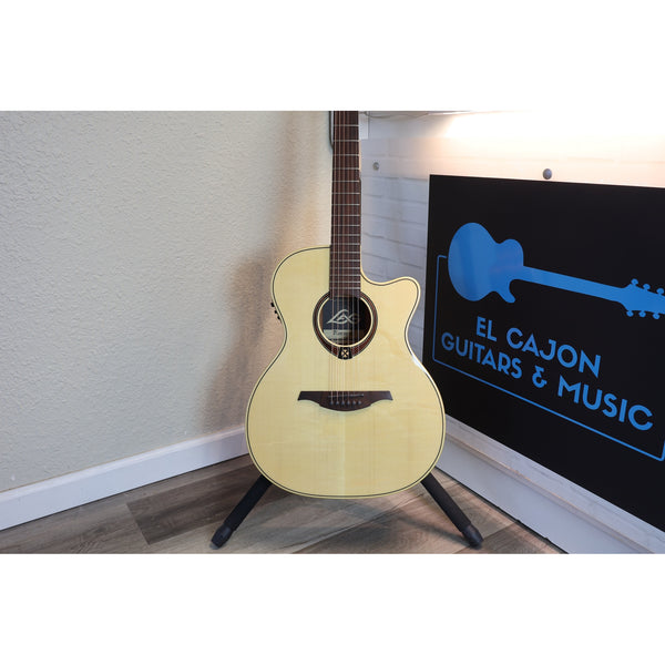 An acoustic cutaway guitar with a natural light wood finish stands on a black tripod in a room corner, next to a black sign with blue text reading 'EL CAJON GUITARS & MUSIC'.