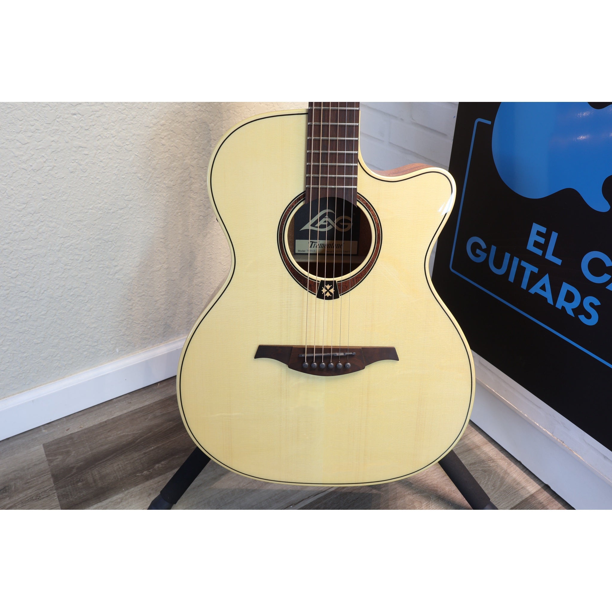 An acoustic cutaway guitar with a natural light wood finish and dark brown neck, displayed on a stand in front of a wall with a sign that reads 'EL C GUITARS'.