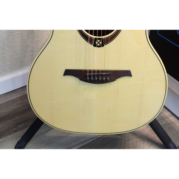 Close-up of an acoustic guitar body on a stand, showing a light natural wood finish, dark bridge, and part of the soundhole rosette in an indoor setting.