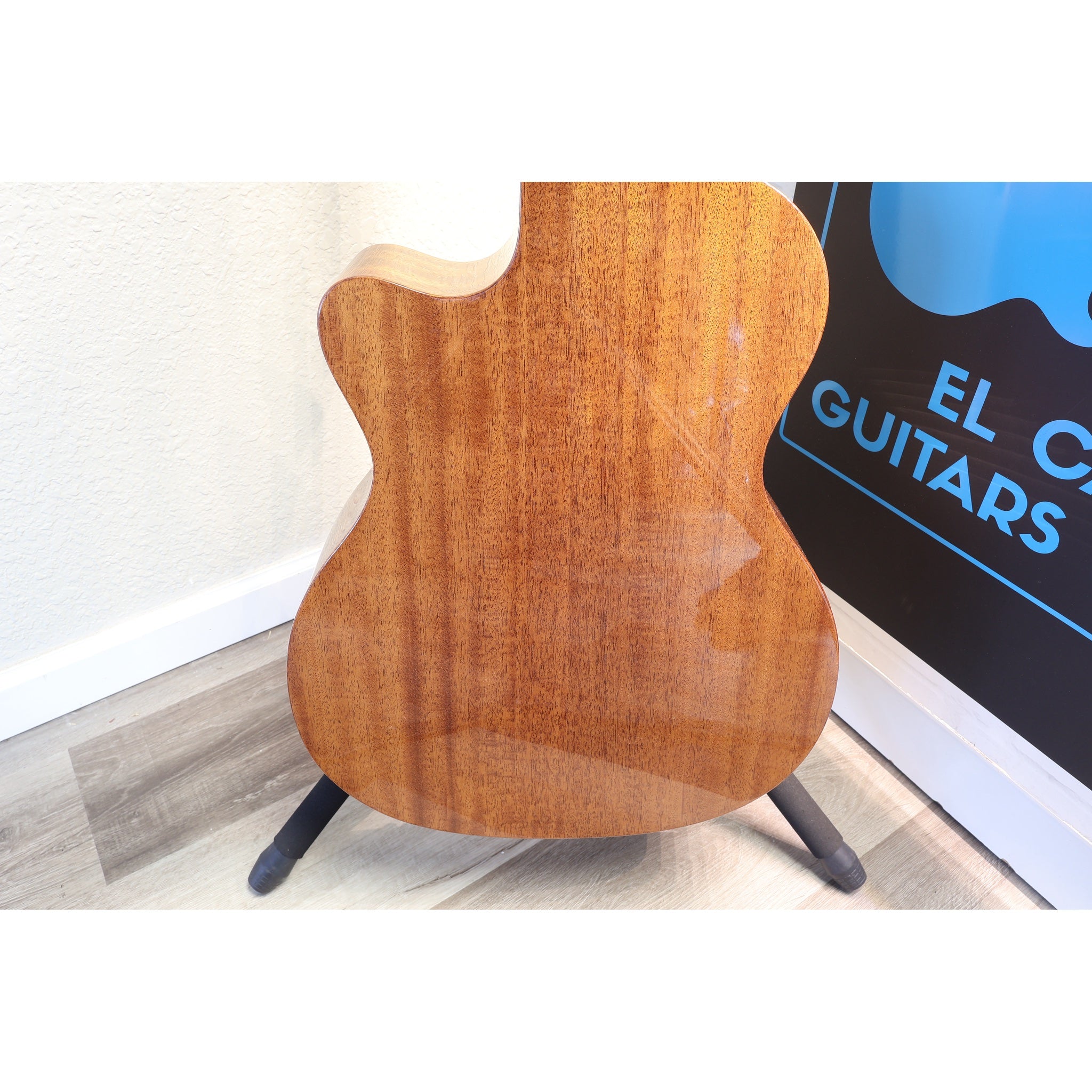 The back of a wooden electric guitar with a glossy finish, displayed on a black stand on a light wood floor next to a wall and a sign that reads 'EL C.