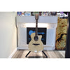 An acoustic cutaway guitar with a natural wood finish stands on a black tripod stand in front of a white brick wall display.