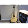 A natural wood acoustic guitar with a cutaway body stands on a black metal stand in an indoor room. A piano is visible on the right, along with a sign that reads 'B.