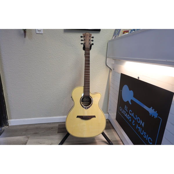 A natural wood acoustic guitar with a cutaway body stands on a black metal stand in an indoor room. A piano is visible on the right, along with a sign that reads 'B.