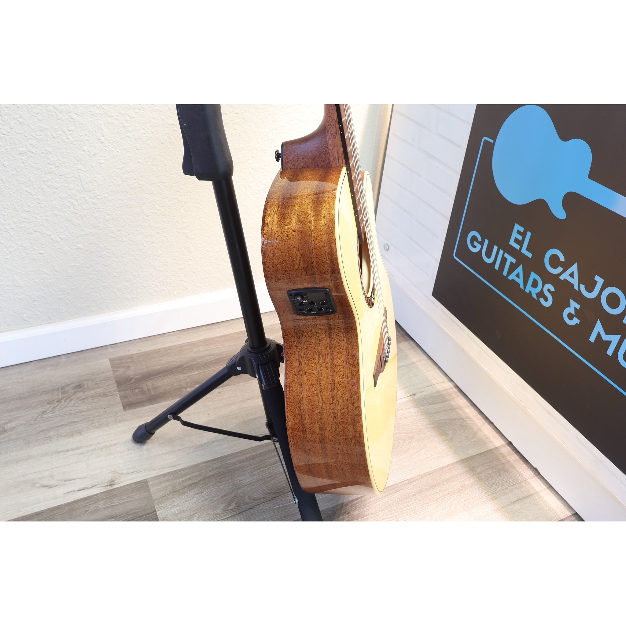 An acoustic guitar with a natural wood finish stands on a black music stand in an indoor room with a light-colored wall and wood-look floor. A sign reading 'EL CAJON GUITARS & M.