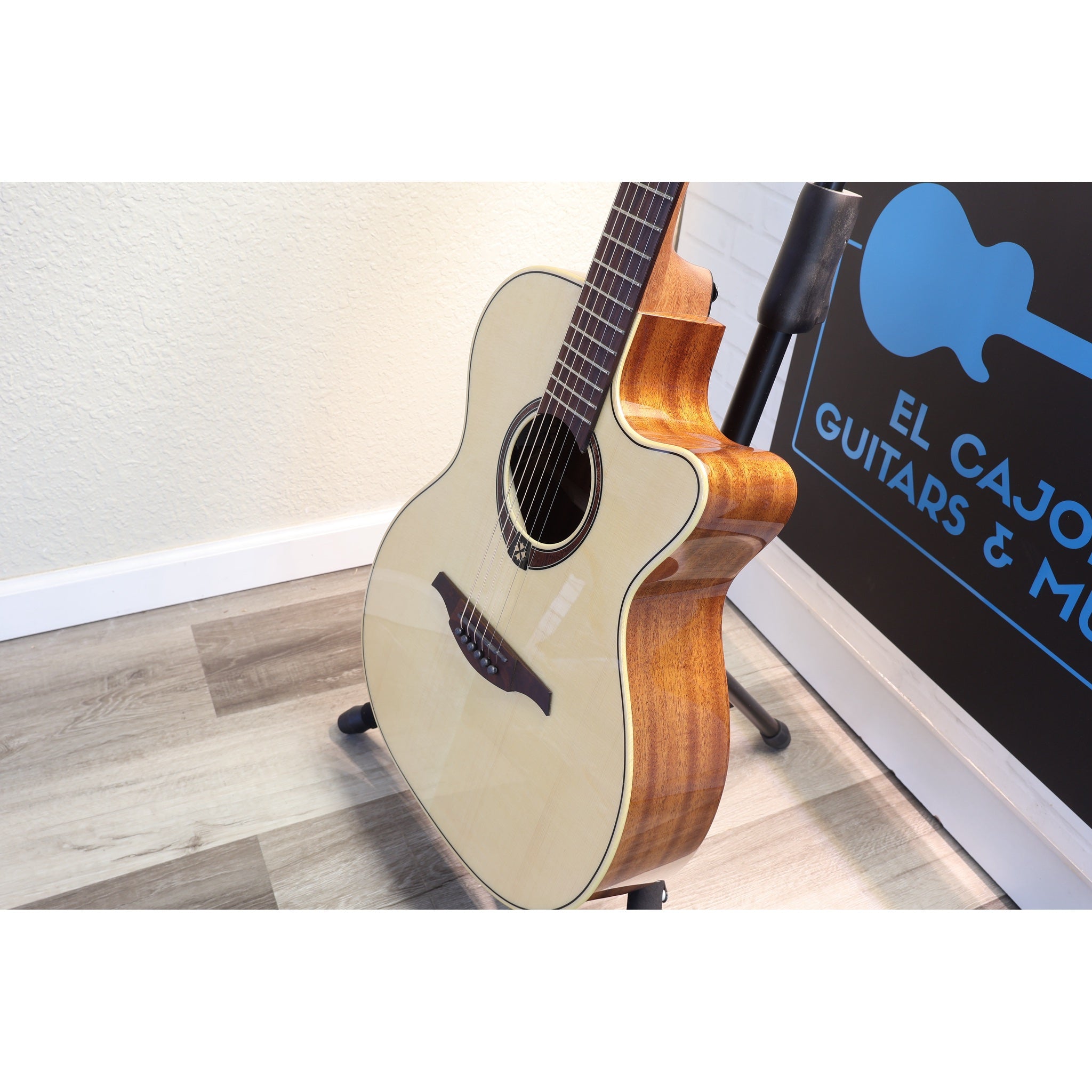 An acoustic cutaway guitar with a natural wood body and cream-colored top, displayed on a stand in an indoor setting next to a sign that reads 'EL CAJON GUITARS & MUSICAL INSTRUMENTS'.
