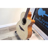 An acoustic cutaway guitar with a natural wood body and cream-colored top, displayed on a stand in an indoor setting next to a sign that reads 'EL CAJON GUITARS & MUSICAL INSTRUMENTS'.