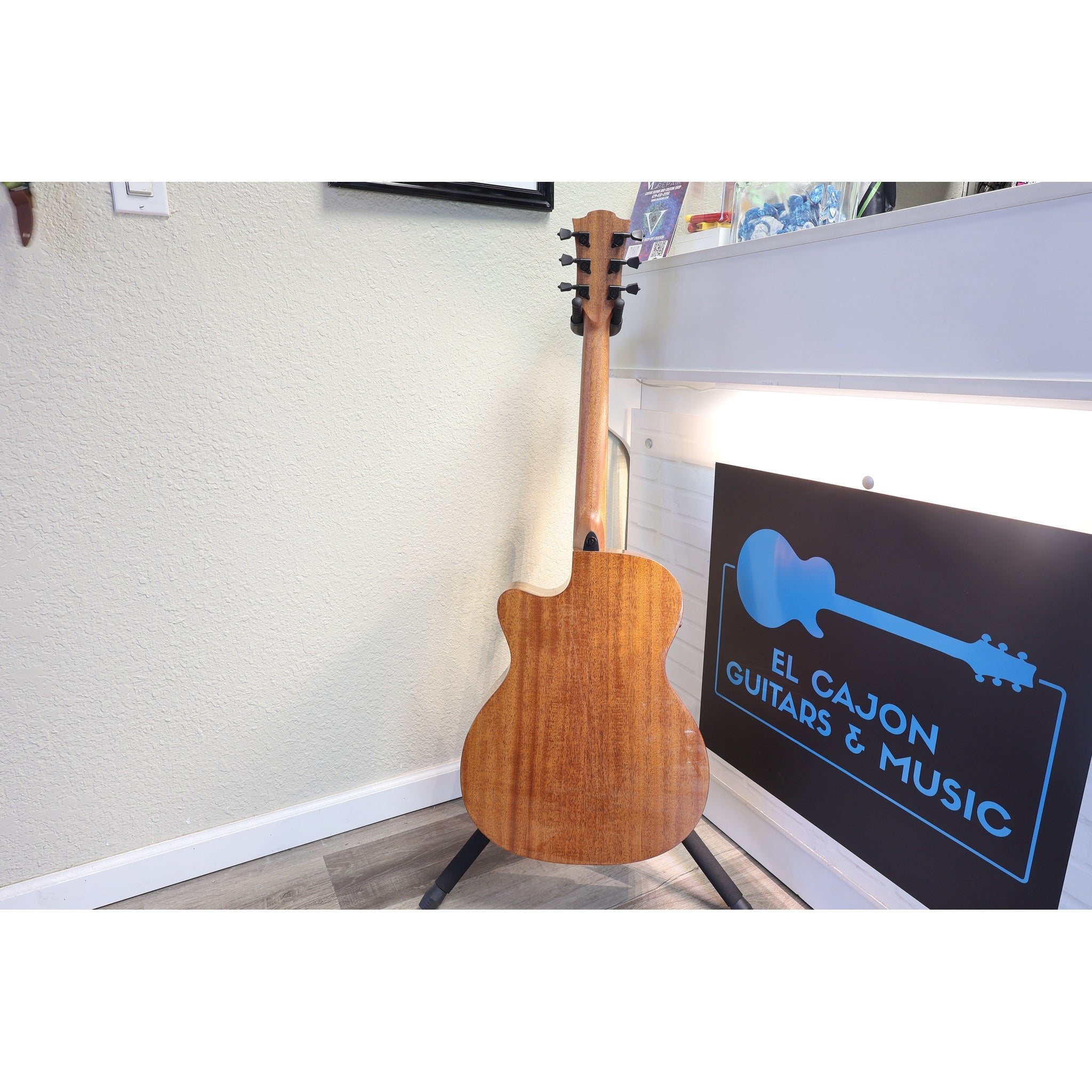 An acoustic guitar with a natural wood finish stands on a black stand in an indoor room. A sign on the right reads 'EL CAJON GUITARS & MUSIC'.