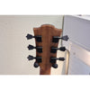 Close-up of the headstock of a wooden stringed instrument, likely a guitar or ukulele, showing six black tuning pegs against a light wall background.